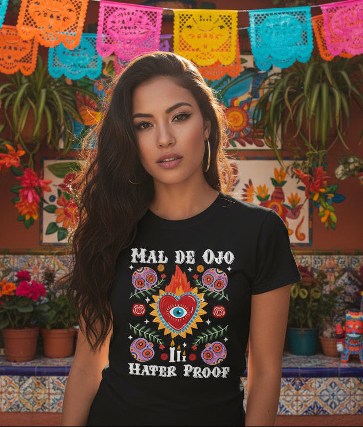 Black t-shirt featuring a colorful sacred heart with a blue eye in the center, sugar skulls, and the phrase Mal de Ojo Hater Proof.